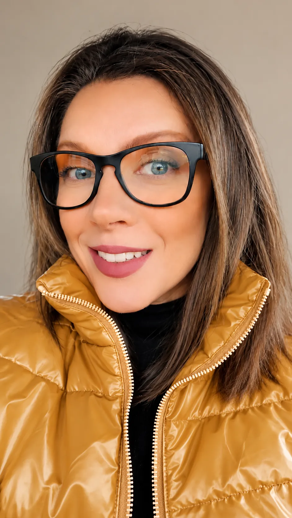 Luciana Adam sorrindo, de óculos, colete mostarda e gola alta preta.